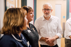 Eine Gruppe von Menschen, die in einem Raum mit einer Tür im Hintergrund und einem Anschlagbrett mit Papierschnipseln darauf auf der linken Seite stehen und sprechen.