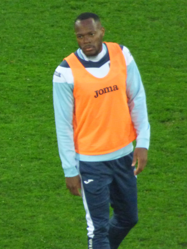 Ein Fußballer in einem orangen T-Shirt und blauen Hosen geht entschlossen über ein grünes Feld.