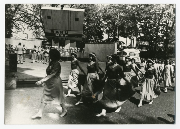 Eine Gruppe von Frauen, die auf der Straße tanzen, umgeben von einer Menschenmenge, einem Lautsprecher, einem Fahrzeug, einer Tafel mit Text und einer Fahnenstange.