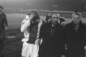 Eine Gruppe von Männern in Not steht zusammen auf einem Fußballfeld, mit einem Stadion im Hintergrund, in einem Schwarz-Weiß-Bild.