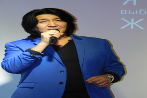 Ein Mann in einem blauen Anzug und einem schwarzen Hemd hält ein Mikrofon und steht vor einem Bildschirm mit Text, mit einem ernsten Gesichtsausdruck.