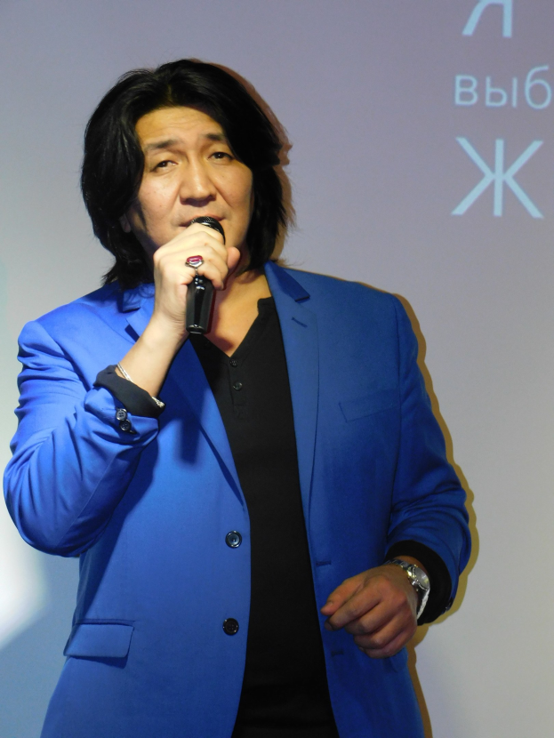 Ein Mann in einem blauen Anzug und einem schwarzen Hemd hält ein Mikrofon und steht vor einem Bildschirm mit Text, mit einem ernsten Gesichtsausdruck.