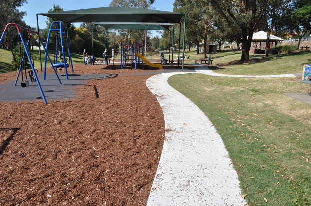 Ein Park mit einem Spielplatz, der Schaukeln und Rutschen bietet, umgeben von grünem Gras, Bäumen, Bänken, Schuppen im Hintergrund und einem klaren blauen Himmel.