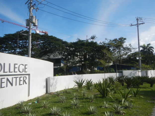 Eingang zu einem College-Zentrum mit einem "College-Zentrum"-Schild an der Wand, umgeben von Grün, einem Weg, Strommasten, Bäumen, Gebäuden, einem Kran und einem bewölkten Himmel.