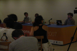 Eine Gruppe von Menschen, die auf Stühlen vor einem Tisch mit einem Mikrofon, einem Laptop, Flaschen und einer Kamera sitzen, mit einer Wand im Hintergrund.