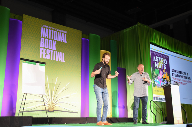Zwei Männer auf einer Bühne beim Nationalen Buchfestival, mit einem großen Bildschirm dahinter und einem Rednerpult mit Mikrofon auf der rechten Seite.