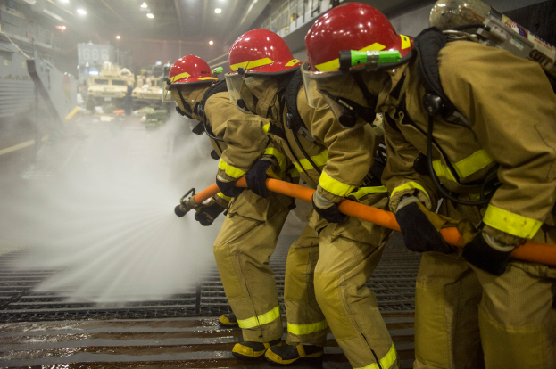 Feuerwehrleute in Helmen und Handschuhen sprühen Wasser aus Rohren auf ein Feuerwehrauto, mit Lampen, Geländern und Texttafeln im Hintergrund.