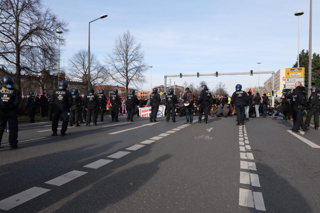 Eine Gruppe von Polizisten in schwarzen Uniformen und Helmen, die auf der Seite einer Straße mit Laternen, Ampeln, Bäumen, Gebäuden und einem klaren blauen Himmel im Hintergrund stehen.