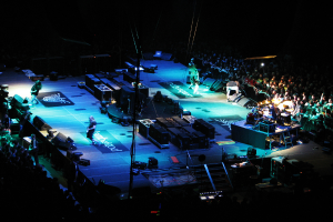 Eine Gruppe von Menschen, die auf einer Bühne mit Lautsprechern, Lichtern und Mikrofonen performen, umgeben von einer großen Menge in einem dunklen Hintergrund.