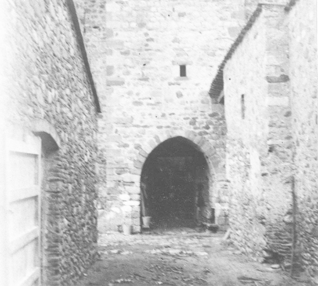 Schwarzes und weißes Foto eines alten Steingebäudes mit einem zentralen Torbogen, flankiert von Mauern, beschriftet mit "Der Eingang zur Burg" unten.