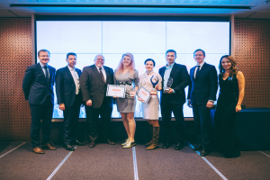 Eine Gruppe strahlender Menschen hält Zertifikate und steht zusammen vor einem Bildschirm, mit einem rechts daneben stehenden, mit schwarzem Tuch bedeckten Tisch und einem Gegenstand an der Wand im Hintergrund, feiert ihren Sieg bei den Internationalen Business Awards 2018.
