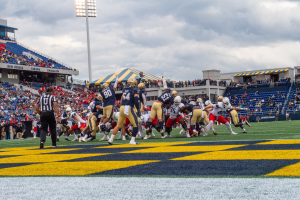 Ein Fussballspiel im Gange mit Spielern in Sportbekleidung und Helmen im Vordergrund, Zuschauern im Hintergrund und Stadionelementen wie Stühlen, Geländern, Schildern, Leuchten und Text unten.