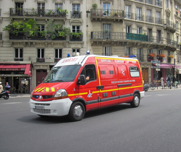 Ambulanz auf dem Seitenstreifen vor einem Gebäude mit Fenstern, Geländern, Pflanzen und anderen Gegenständen geparkt, mit einigen Menschen und Fahrrädern im Hintergrund.