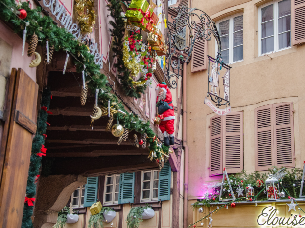 Ein Gebäude in Colmar, Frankreich, geschmückt mit Weihnachtsdekorationen, darunter Girlanden, Lichter und eine Santa-Claus-Figur, mit einem Schild, auf dem "Colmar Weihnachtsmarkt" steht.