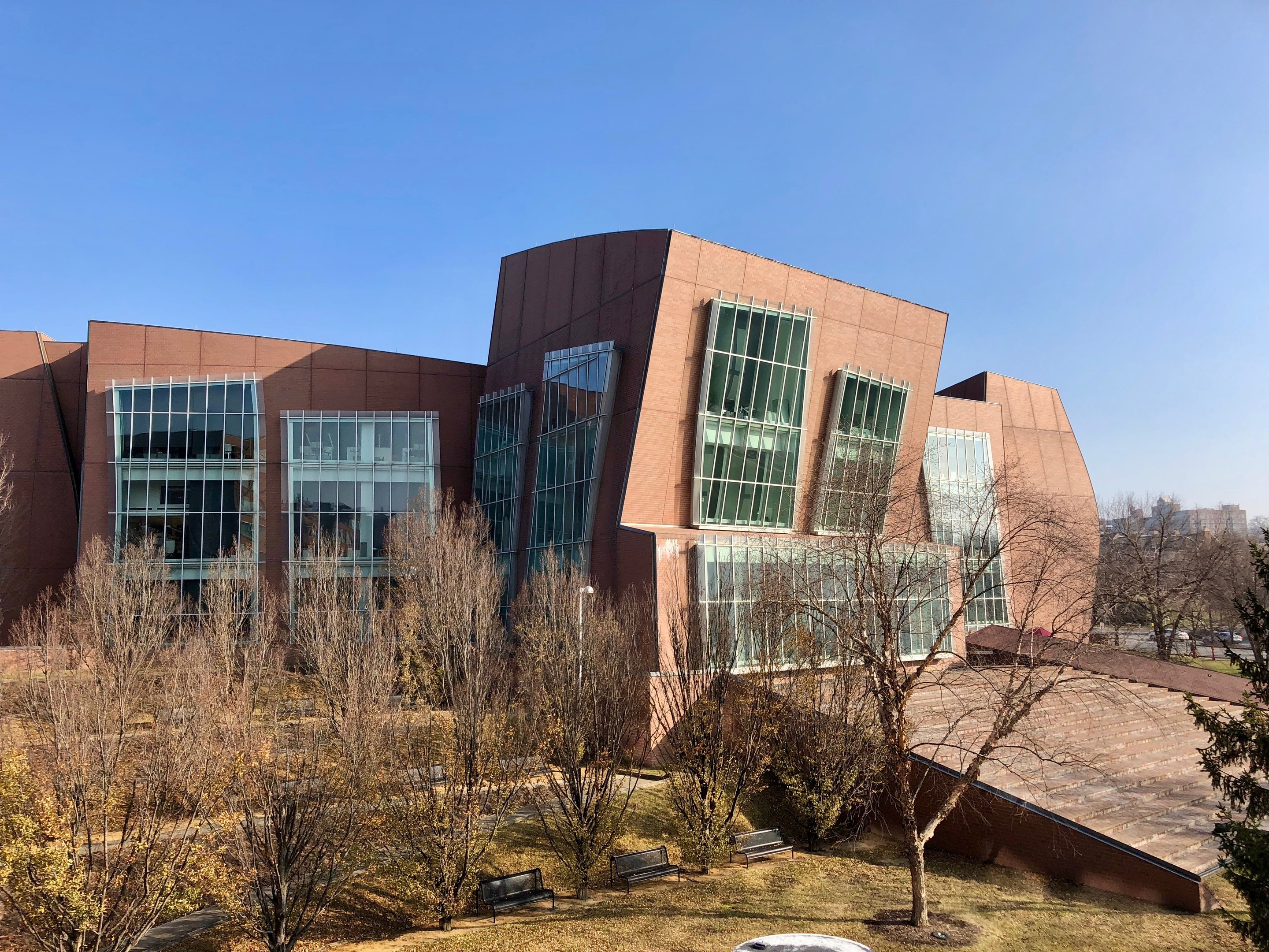 Großes Gebäude mit vielen Fenstern, umgeben von Bäumen, Bänken, Gras und einem Laternenmast, vor anderen Gebäuden und einem klaren blauen Himmel, als der neue Campus der University of Illinois at Chicago identifiziert.