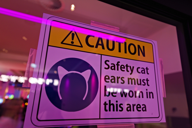 Hellgelbes Warnschild in einem Schaufenster mit der Aufschrift "Vorsicht: Sicherheits-Katzenohren müssen in diesem Bereich getragen werden" vor einem unscharfen Hintergrund.