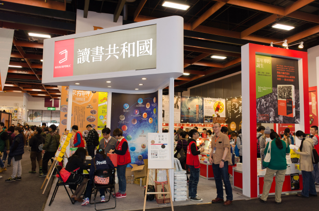 Eine große Gruppe von Menschen an einem Messestand mit Text "Book Republic Hong Kong Book Fair", Bannern und Deckenbeleuchtung.