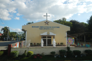 Eine unabhängige Kirche mit einem Kreuz vorne drauf, umgeben von einem Zaun, Statuen, Pflanzen, Gras, einem Weg, Bäumen, Pfählen, Drähten, einem nahen Haus und einem bewölkten Himmel.