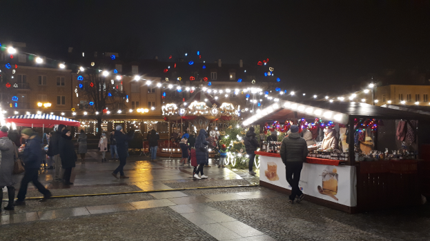Ein lebendiger Weihnachtsmarkt in einer Stadt bei Nacht, mit Menschen, die herumlaufen, Ständen mit Lichtern und Dekorationen, Gebäuden mit Fenstern, Bäumen und einem Himmel, der mit festlichen Lichtern erleuchtet ist.