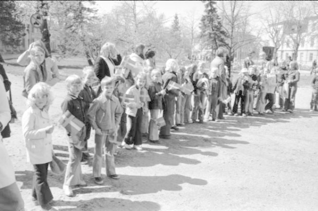 Schwarz-weißes Bild einer Gruppe von Menschen, die Fahnen in einer Reihe auf einem Schotterweg während einer Protestmarsch auf dem Schulgelände halten, mit Bäumen, Gebäuden und einem klaren Himmel im Hintergrund.