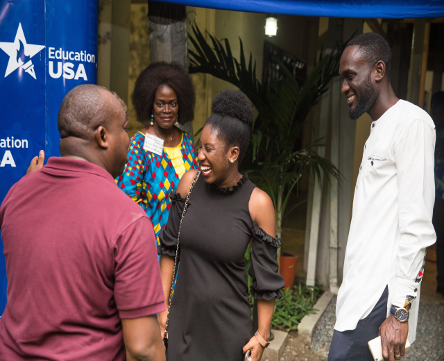 Gruppe von Menschen, die vor einem blauen Zelt mit einer Fahne, Topfpflanzen, einer Säule, einer Wand und einer Decke mit Leuchten sprechen.