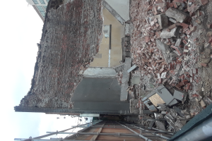 Ein teilweise abgerissener Bau mit verstreuten Ziegeln und Metallstangen, umgeben von anderen Gebäuden unter einem klaren blauen Himmel.