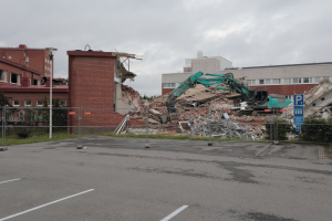 Großes Gebäude in einer Parkanlage mit einem Bagger, Metallstangen, Zaun, Hinweisschild, Pflanzen, Gras, Bäumen, benachbarten Gebäuden und einer bewölkten Himmel.