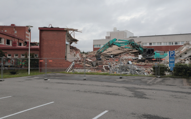 Großes Gebäude in einer Parkanlage mit einem Bagger, Metallstangen, Zaun, Hinweisschild, Pflanzen, Gras, Bäumen, benachbarten Gebäuden und einer bewölkten Himmel.