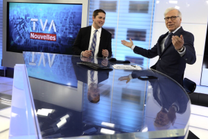 Zwei Männer in Blazern, Krawatten und Brillen sitzen an einem Glastisch in einem Nachrichtenstudio, mit einer Tastatur, einer Maus, Papieren und einem Handy auf dem Tisch und einem Fernseher und Fenstern im Hintergrund.