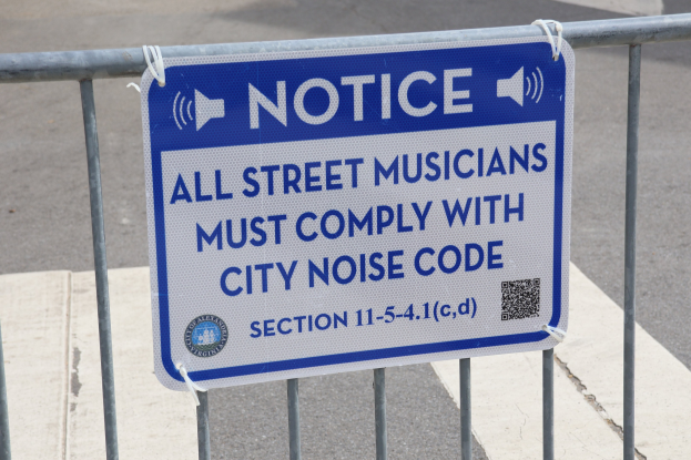 Anschlagtafel an einem Zaun mit einem Schild mit der Aufschrift "Anschlag: Alle Straßenmusiker müssen sich an die städtische Lärmvorschrift halten" und einer Straße im Hintergrund.