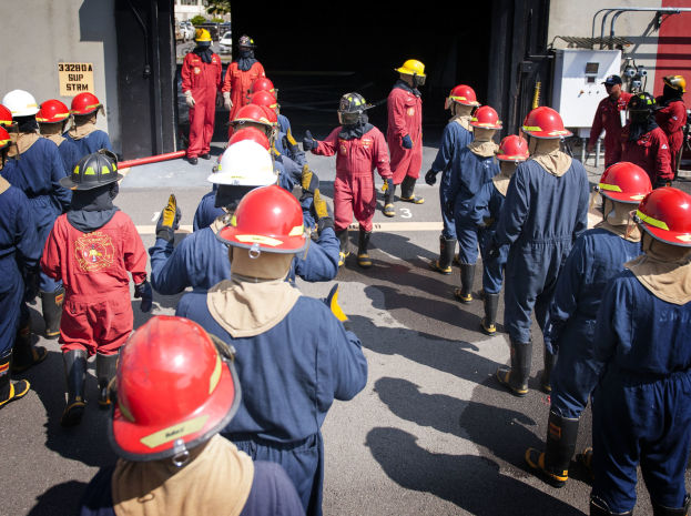 Feuerwehrleute in roter Uniform und Helmen gehen eine Straße entlang, zu ihrer Linken eine Wand mit Graffiti, Bäume und einen klaren blauen Himmel im Hintergrund.