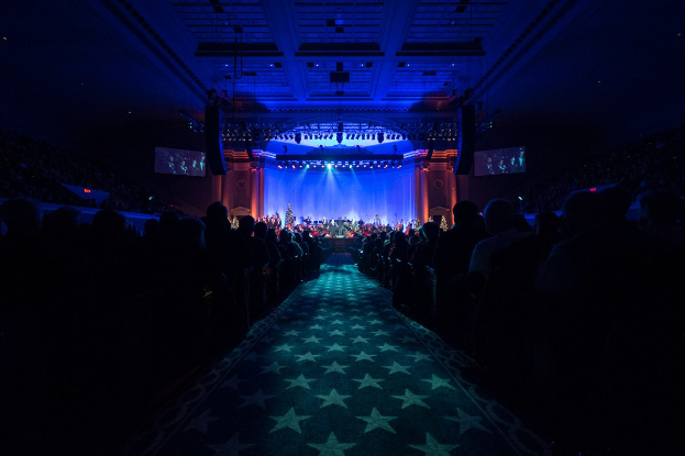 Ein großer Auditorium mit sitzenden Zuschauern, eine Bühne mit Musikern und Deckenbeleuchtung und -fernseher, was auf ein Konzerthaus während der Nationalkonvention 2019 hinweist.