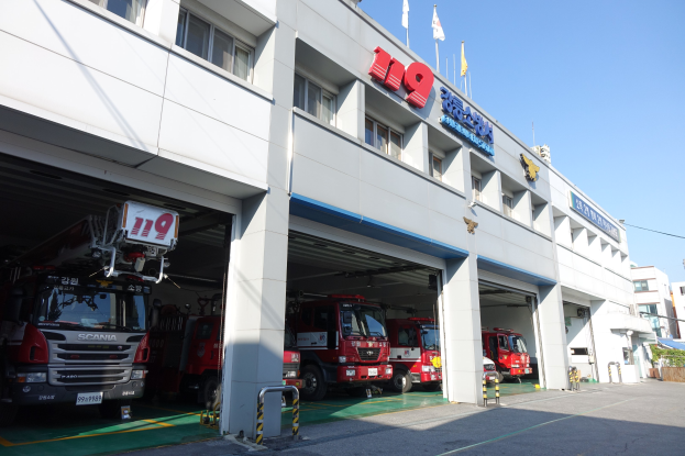 Feuerwache mit mehreren Feuerwehrautos vor der Tür, mit Fenstern, Säulen, Namensschildern und Flaggen im Vordergrund; andere Gebäude und ein klarer blauer Himmel im Hintergrund.