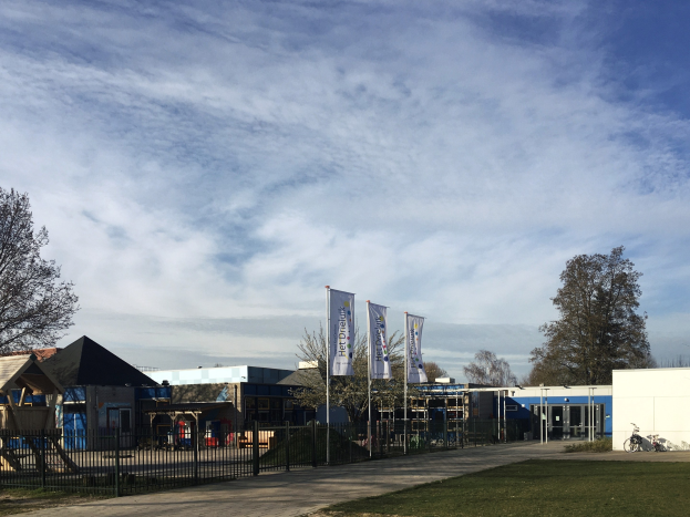 Großes Schulgebäude mit einem Kinderspielplatz davor, umgeben von einem Metallzaun, Gras, Bäumen, einem Weg, Bannern, einem geparkten Fahrrad, nahen Häusern und einem bewölkten Himmel.