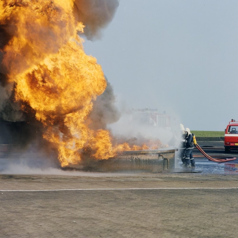 Feuerwehrlöschfahrzeug in Flammen auf der Stra√enseite mit zwei Helmen tragenden Individuen mit Schläuchen, einem Fahrzeug im Hintergrund und dem Himmel.
