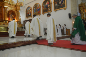 Eine Gruppe von Priestern geht einen roten Teppich in einer Kirche hinunter, mit Menschen, die auf dem Boden vor und rechts von ihnen stehen, und Foto Rahmen an der Hintergrundwand.