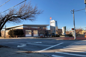 Großes Gebäude mit einer Flagge auf dem Dach, identifiziert als neue Feuerwache an einer Straßenkreuzung, umgeben von Bäumen, Straßenmöbeln und Infrastruktur, unter einem sichtbaren Himmel.