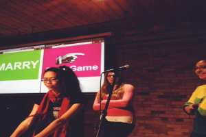 Drei Frauen stehen mit dem Gesicht nach links, mit einem Mikrofonständer in der Mitte und einem Bildschirm mit Text auf der linken Seite.