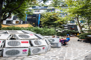 Luftkühler am Boden links unten, eine Straße nach rechts, Menschen auf Stühlen neben der Straße, mit Gebäuden, Bäumen und Pflanzen oben.