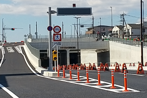 Eine Stra√üe mit orangen Verkehrskegeln am Rand, eine Br√ľcke im Hintergrund, Laternen, Ampeln, Verkehrszeichen, Stromm√§ste mit Dr√§hten, Geb√§ude und ein bew√∂lkter Himmel.