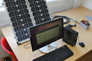 Ein Computersystem mit Tastatur, Maus und CPU auf einem Tisch, neben einer Solarzelle, einer Batterie, Chips und Drähten, mit einem roten Stuhl und einer weißen Wand im Hintergrund.