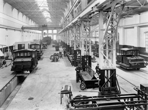 Schwarzes und weißes Foto einer Werkhalle mit mehreren Bahnen unter Reparatur, Arbeitern, die sich darum kümmern, Metallpfosten, die die Struktur stützen, Deckenlampen und Fenstern an den Wänden.