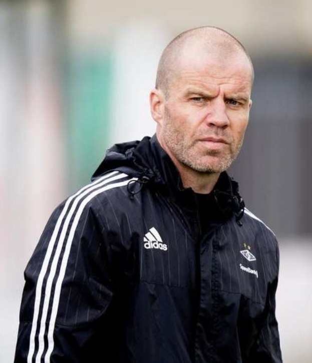 Ein kahlköpfiger Mann in einer schwarzen Jacke, der offenbar ein Fußballtrainer ist, steht auf einem Feld und hat die Hände gefaltet und einen ernsten Gesichtsausdruck. Er trägt eine blaue Jacke mit weißen Streifen und einem weißen Kragen.