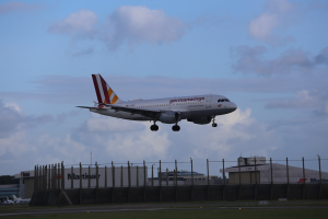 Germanwings Airbus A320-200 beim Start von Frankfurt Airport, mit Gras darunter, einem Zaun im Vordergrund, Gebäuden und Bäumen im Hintergrund und einer bewölkten Himmel über