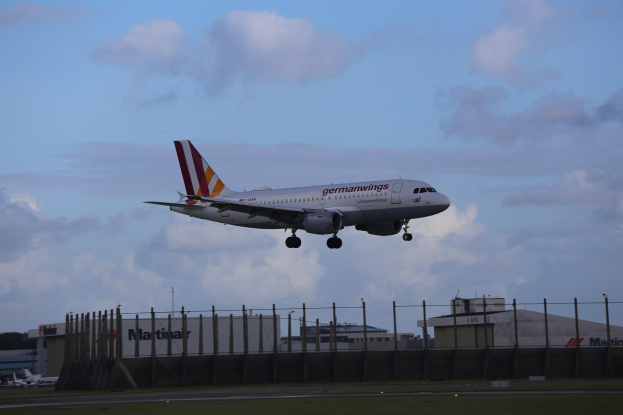 Germanwings Airbus A320-200 beim Start von Frankfurt Airport, mit Gras darunter, einem Zaun im Vordergrund, Gebäuden und Bäumen im Hintergrund und einer bewölkten Himmel über