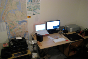 Ein Raum mit einem Tisch, auf dem ein Laptop, ein Lautsprecher, ein Monitor, eine Tastatur, ein Ball, eine Maus, ein Scanner und Papiere stehen; ein weiterer Tisch mit elektronischen Geräten ist links unten zu sehen, mit Charts und Postern an der Wand dahinter.