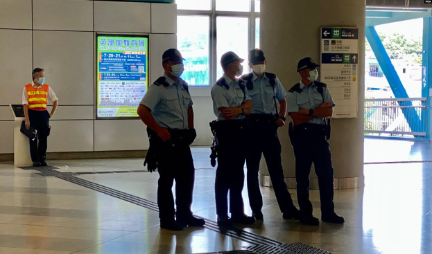 Gruppe von maskierten Polizeibeamten vor einem Gebäude mit Glastüren, textbedeckten Tafeln, einer Säule und einem Geländer, mit Bäumen und Fahrzeugen im Hintergrund durch die Glasscheibe sichtbar.