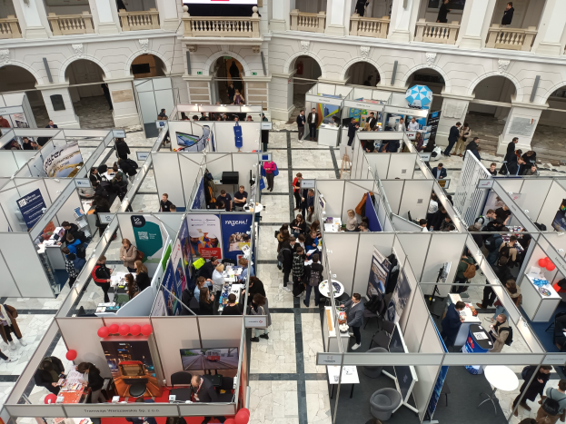 Große Gruppe von Menschen auf einer internationalen Messe mit Ständen, Bannern, Tischen, Stühlen, Luftballons und einem Gebäude mit Bögen, Säulen, Geländern und einem Hintergrundbild.