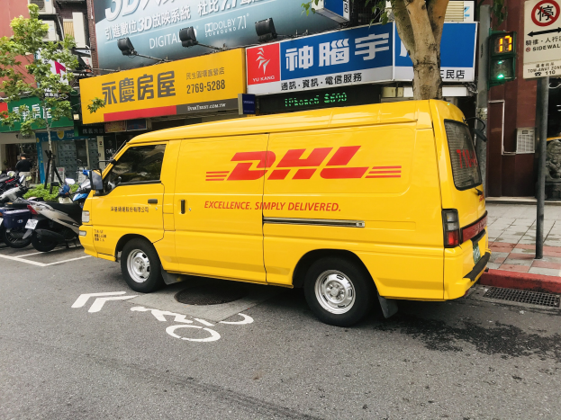 Ein gelber DHL-Lieferwagen parkt auf einer Straße neben Gebäuden, Bäumen, Schildern, Masten, Verkehrszeichen und einem Fussgänger auf dem Gehweg.