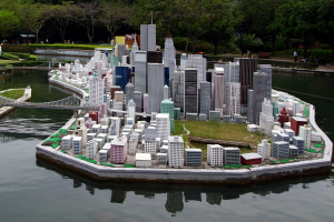 Ein detailliertes Miniaturmodell einer Stadtlandschaft, umgeben von Wasser, mit Gebäuden, Grünflächen, einer Brücke, Menschen, Pflanzen, Bäumen und einem Himmelhintergrund.